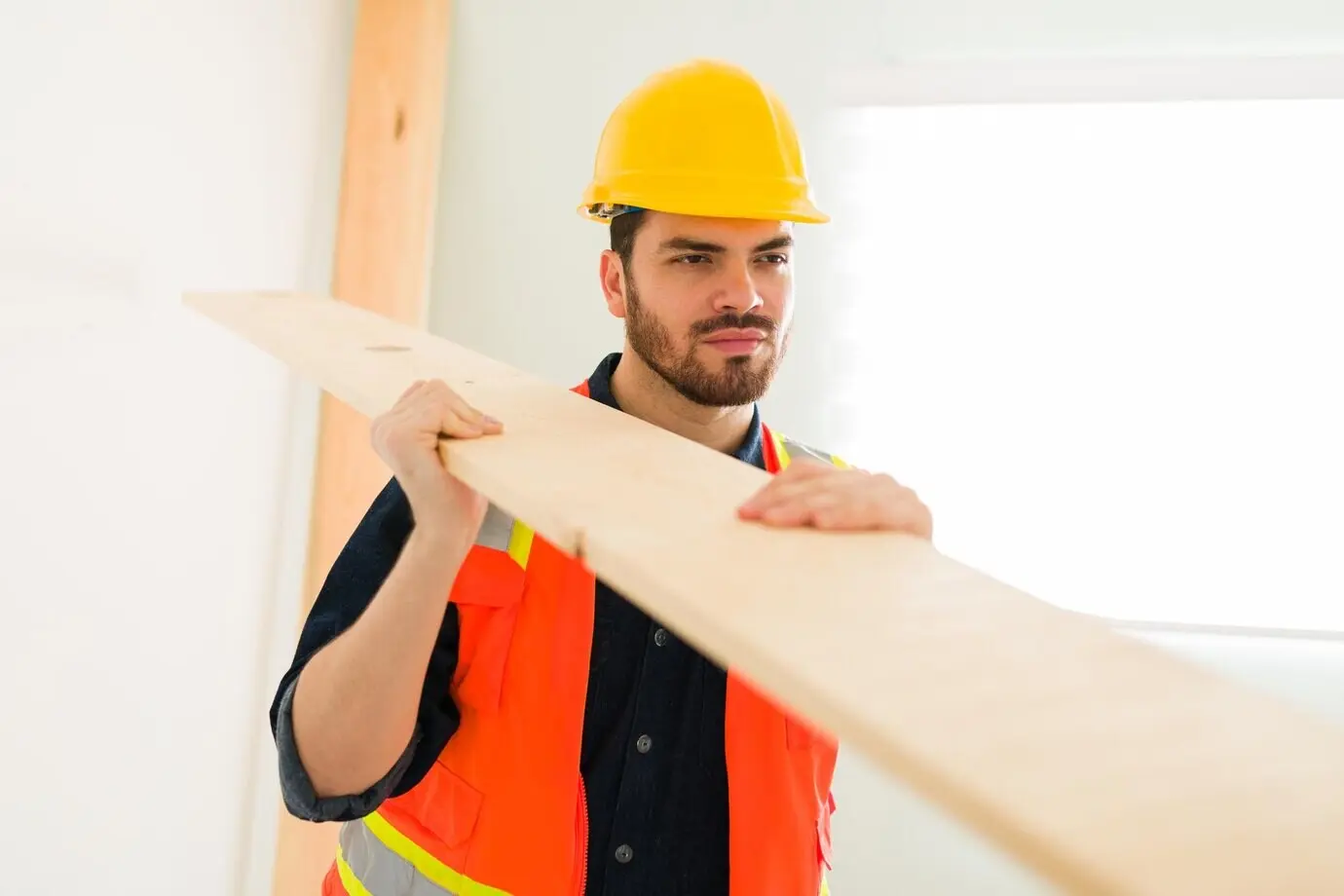 A handsome Latin engineer wearing a helmet and safety vest carries a wooden panel at the construction site.